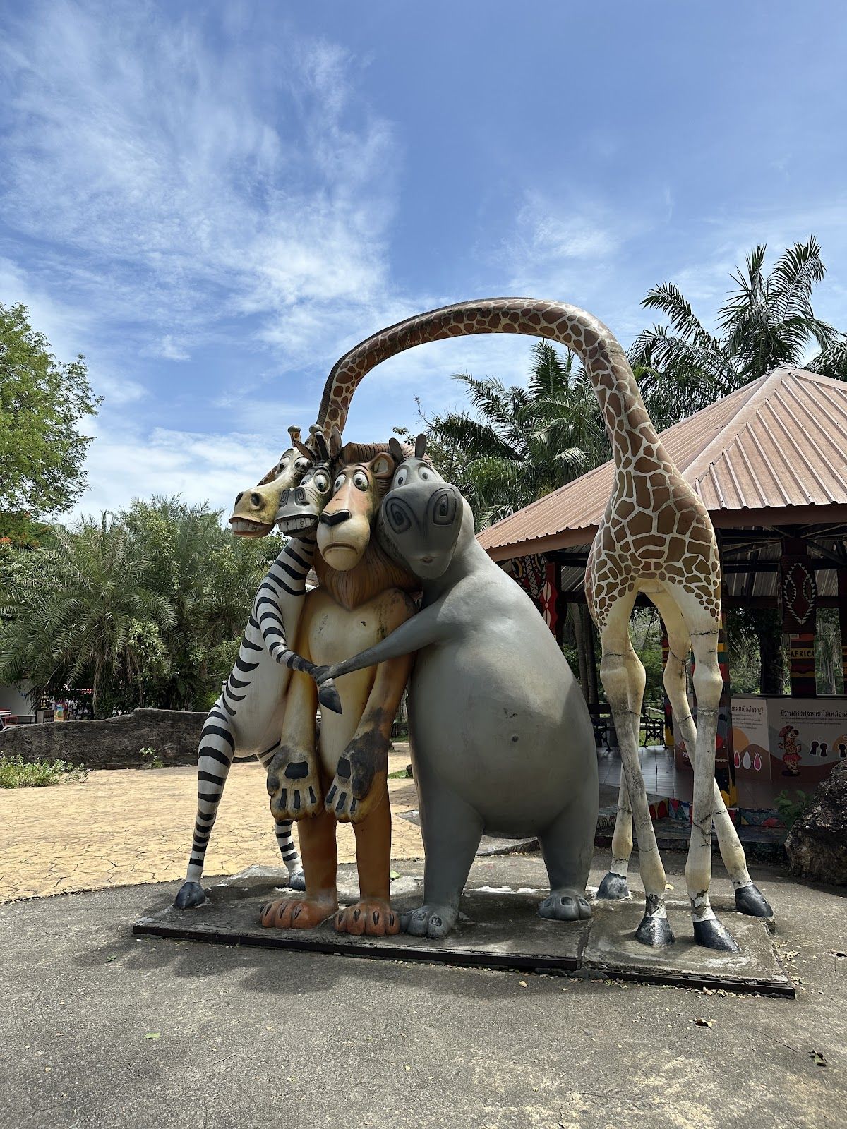 A visitor hand-feeding a giraffe that leans its long neck over the fence at Khao Kheow Open Zoo, with green trees and sandy enclosure behind